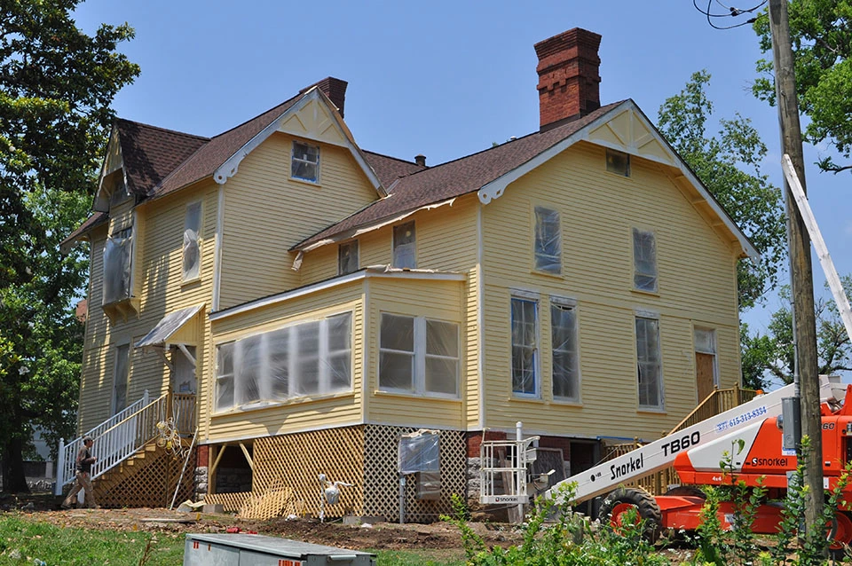 House with white siding, windows boarded up and construction taking place.