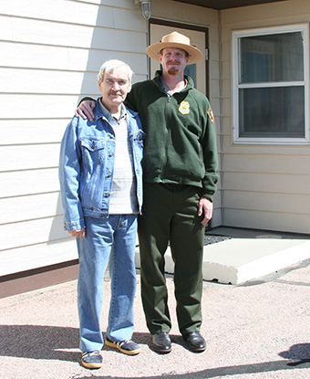 Older man and a park ranger stand in front of a tan building.
