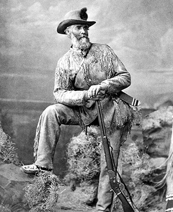 Norris wearing a frilled leather jacket and pants, a hat, and leaning against a rifle.