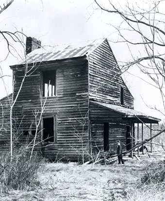 black and white photo of a two story wooden house with a tall thin tree in the left side of the yard