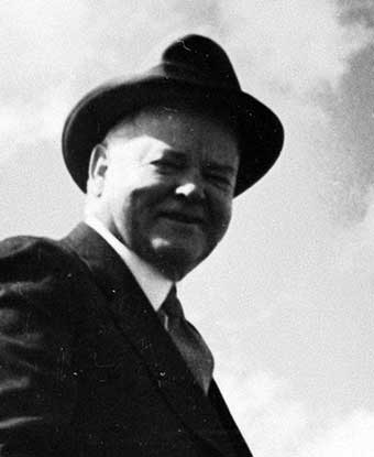 A black and white photo depicts a smiling middle aged man in a hat outdoors. A black and white photo depicts a smiling middle aged man in a hat outdoors.