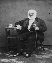 Elderly man in a suit sitting in a chair with a walking cane.