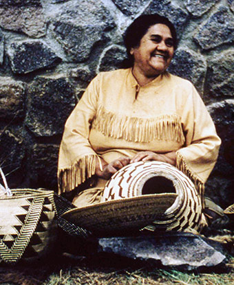 A smiling woman in American Indian attire sits with woven baskets