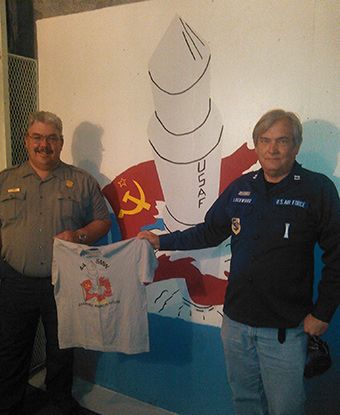 Two men hold a t-shirt in front of a wall painted with a design identical to the shirt.