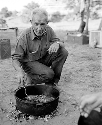 A man kneels next to a dutch oven on the ground.