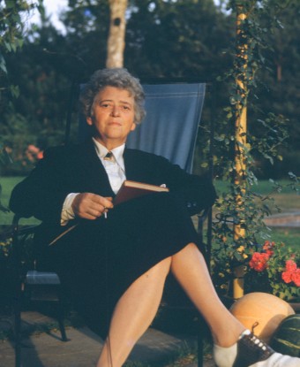 A woman seated outdoors with a book in her hand and wearing a dark suit.