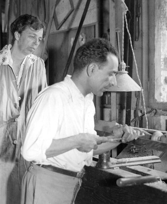 A woman looks over the shoulder of a man carving wood on a lathe.