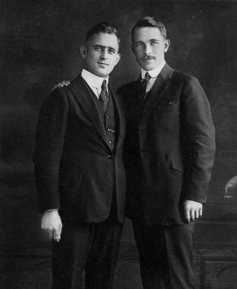 Two men in dark suits embrace one another's shoulder looking toward the camera.