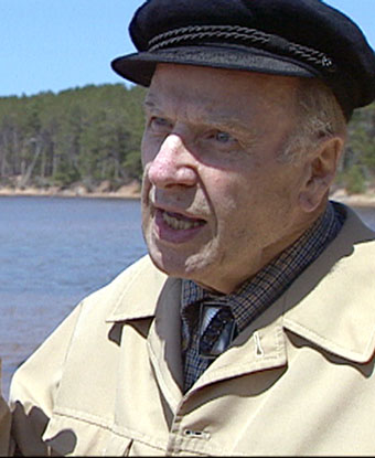 An older man with a black hat and khaki jacket with water and trees in the background. 