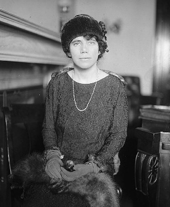 A seated woman wearing a fur hat, pearls, and with fur stole across her lap.