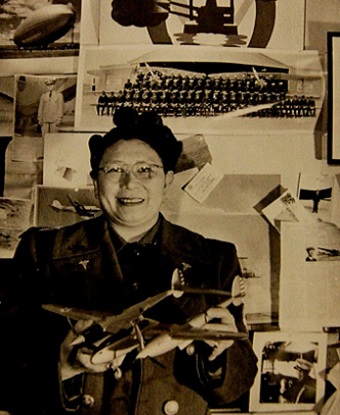 Woman wearing glasses and holding model airplane, in front of photos of military servicemen.