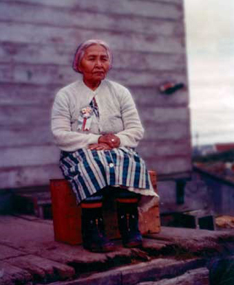 Image of a woman sitting on a wooden box. She is squinting and her hands are crossed in her lap.