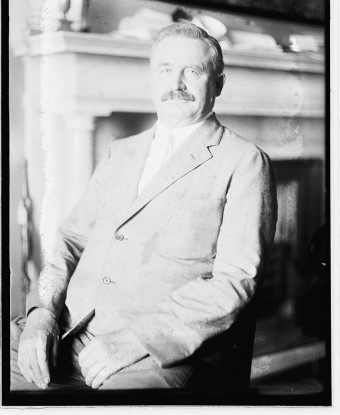 a mustachioed man in a suit smiles as he sits in front of a fireplace.