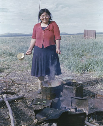 A woman stands before a steaming pot sitting in the dirt. She is holding a pot and is smiling.