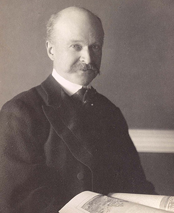 A suited, three-quarter pose of a balding man with mustache, holding an open book in his lap.