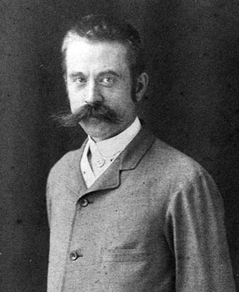 Head and shoulders portrait of a man wearing a suit. He has a large mustache and intense gaze.