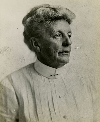 Black and white photo of an older woman, hair in a bun, pleated white shirt pinned at the collar.