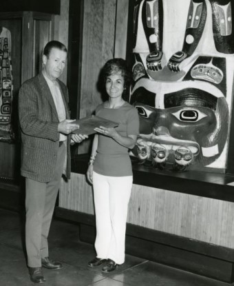 A woman and man smile at the camera. There are pieces of Alaskan Native artwork on wall behind them