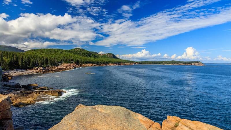 Rocky Maine coast on left with bright blue ocean on right. Coast line seen in the distance covered in large evergreen trees.