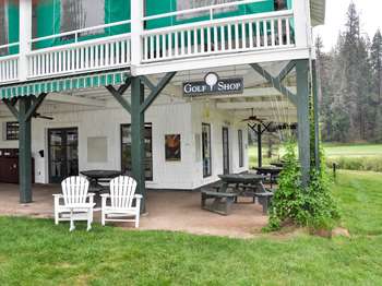 A sign that reads, 'Golf Shop,' hangs from the ceiling at the corner of a two story building. Circular picnic tables are beneath a porch and lawn chairs sit on the green grass that surrounds the building.