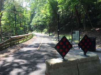 A paved road curves out of forested area. Two cement blocks prevents cars from driving into a pullout