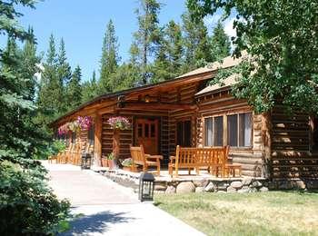 Approach to Jenny Lake Lodge front door. Brown log cabin with wooden chairs on walkway and small patio. Flower baskets hang from eaves.