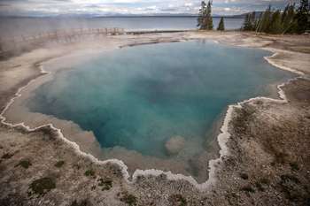 A large hot spring steaming in front of a large lake, with a wooden boardwalk going between them.