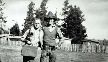 Two men in Western garb stand, arm in arm, in front of rustic cabins and fencing.