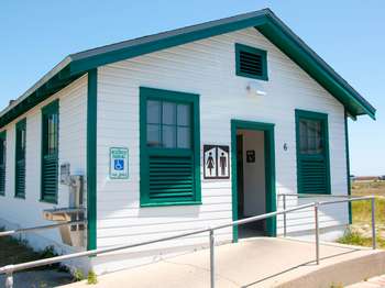 A white building with green trim. On the building hangs a brown and white restroom sign and a handicap parking sign. A cement walkway with metal railings in front of the building. Blue sky in the background.