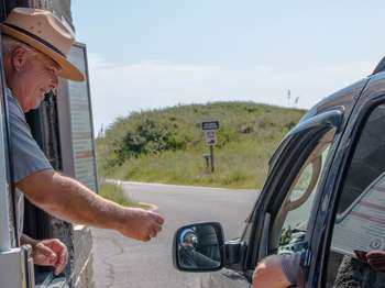 A park ranger wearing a tan hat leans out of a grey stone building on the left. A black stopped car with a person's elbow sticking out the driver's window on the right. Green grass on a sand dune and a bright blue sky in the background.