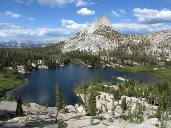 A dual pointed peak rises above a surrounding forest near a lake. Puffy clouds fill the sky.