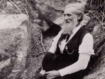 A black and white photo shows an elderly man sitting on a boulder. He white hair and a long beard.