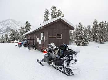 Snow-covered, wooden brown warming hut with a snowmobile parked in front of the building.