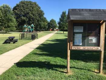 A bulletin board stands beside a sidewalk that leads past a playground and restrooms.