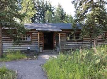 Maud Noble cabin brown log with white chinking. Spruce trees grow overhead.