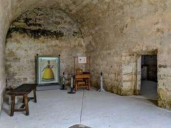 The Second Spanish Room can be accessed from the courtyard, the British room, or the Chapel. This audio description is from the courtyard entrance. This room shows exhibits, in English and Spanish, describing the Castillo at the time that the Spanish returned, in 1784. Two wooden doors open inward to the room. Above the door is a barred window with a wooden shutter that also opens inward. Barred windows flank the doorway. An entrance to the British room is on the left, wall and entrance to the Chapel is on the right. The room is about 20 feet wide and 30 feet long, with an arched ceiling about 17 feet high. To the left is a bench. There is a wooden table with a bench left corner of the back wall. Standing at the entrance to the room, moving from left to right, the exhibits include: Antonia Avero. 1717 to 1792. Text explains that, in Spanish Florida, property often passed from mother to daughter. Many women ran business and amassed large holdings in real estate. Avera left three houses behind when she fled to Cuba when the British took over. An illustration shows Avera with dark hair, reaching just below her ears and covered in a net. She wears dangling pearl earrings. She has an angular face with black eyebrows and dark set eyes. She wears a pearl choker around her neck. The top of her dress is white, with a square neck top, and full sleeves with long ruffled lace. There is a red ruffled ribbon design at the front. Below is a full yellow skirt with black trim and white lace at the hem. Along the back wall, and roped off, is a wooden desk with a banner suspended above it on a wooden dowel. Behind the desk is a wooden stool and chair. On the left, the banner reads, The King of Spain wants you to join the Spanish Army. Sign up here and collect your pay of one two escudo gold doubloon. And to the right it reads, Presidio de San Augustin, Recruiting Station, Junior Ranger Program, Ages 15 & Under. A wooden box rests on a wooden stand at the right corner of the back wall. Spain Returns 1784 to 1821 Exhibit. The text explains that Florida was returned to Spain in 1784. Thousands of British residents left St. Augustine, but about 300 decided to stay, pledging their allegiance to Spain and the Catholic Church.