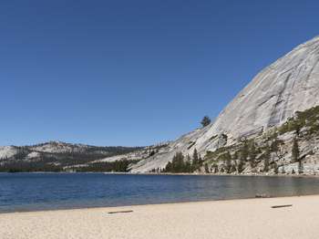 A sandy beach meets the waters' edge of a lake. Large granite domes emerge from the treeline of a surrounding forest.