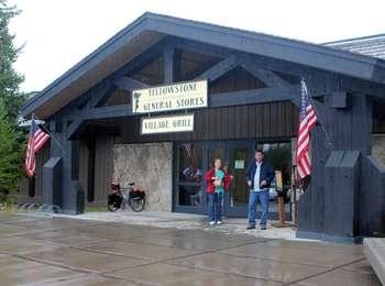 A couple stands between two US flags under the awning of a log entrance under a sign, 