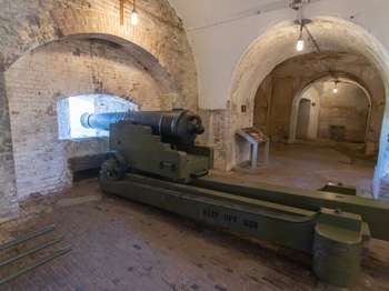 A black metal cannon points out a small window in the white, brick, fort wall. A long hallway of brick arches extends from the right of the cannon.