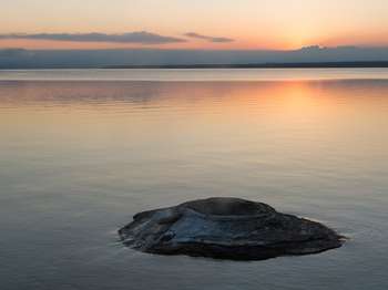 A geyser cone peaks out of the water of a large lake during sunset.
