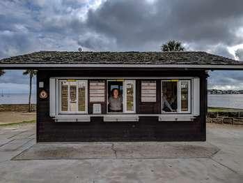 The fee booth, is located at the entrance to the Castillo, at the southeast corner. It is a single-story cabin-style wooden building with a peaked roof. It has dark brown planked wood siding. There are several windows. The south wall has an information bulletin board that reads, Welcome. Castillo de San Marcos National Monument. Hours: 8:45 am to 5:15 pm (last entry at 5 pm). Closed Thanksgiving Day and Christmas Day. Fees: Adults: $15. 15 and Under: Free. A rounded triangle with a white circle with the National Park Service logo, described in more detail later. The words U.S. Fee Area is below the emblem. Text to the right of the triangle reads, Closure. Unmanned Aircraft. Launching, landing or operating an unmanned aircraft (This term includes All types of Devices e.g. Model Airplanes, Quadcopters, Drones, etc.) from or on Lands and Waters Administered by the National Park Service Within the Boundaries of the Castillo de San Marcos National Monument is PROHIBITED. Additional text reads, Your fee dollars at work! Your fee dollars support historic preservation and visitor experience at Castillo e San Marcos National Monument. Some projects include: Ranger Programs. Junior Ranger Activities. Entrance Station Improvements. Castillo Floor Repairs. Covered Way Wall Preservation. Restroom Improvements. Cannon Firing Demonstrations. Text at the bottom of the bulletin reads, Find Your Park A circular logo shows a lake in a grassy area, with three figures near some trees, against a golden sky, with the words, “Every Kid in a Park.â€, The west wall has three windows that slide open, where visitors can pay the entrance fee. Posted on the top center frame is a yellow sign that can slide to read either “openâ€, or “closedâ€,. The glass on the windows has a no smoking sign and signs indicating accepted credit cards, which are VISA, Mastercard, Discover, and American Express. There is a metal ledge at the base of each window. Flanking the center window are Fee Information signs. The U.S. Fee Area logo is posted on the wall, to the left of the windows. The north wall has an information bulletin board, an entry door with a sign that reads “do not block,â€, and a round temperature gauge above the door. The National Park Service logo is shaped like a brown arrowhead, pointed downward. Inside the arrowhead are the words in white lettering, “National Park Serviceâ€, with a green sequoia tree shape to its left. Near the bottom of the arrowhead is a white buffalo silhouette set against a green meadow, with a white lake and snowcapped mountain behind it.