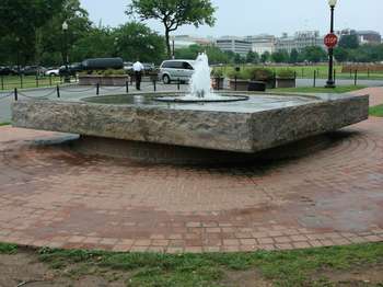 Water sprays up out of the center of fountain that is round pool of water in a stone square base, surrounded by a red brick patio and an urban landscape. 