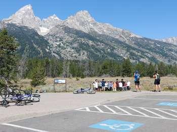 Group of bicyclists at the Teton Glacier turnout sitting on the sidewalk below the high peaks of the Teton Range