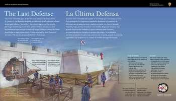 At the top left and center, white text against a blue background in English and Spanish reads:  The Last Defense. The most vulnerable part of the fort is its entrance in front of you. To protect it, the Spanish designed an elaborate set of defenses: a heavy grated gate called a â€œportcullis,â€, 2 drawbridges, and the ravelin. The larger drawbridge took 3 soldiers 15 minutes to close and remained open except in times of danger. Soldiers closed the outer drawbridge at night when most of them returned to their homes in the town. The ravelin protected the fortâ€™s front door.  End of Text  Below, a color illustration of the fort and the drawbridge entrances; text and thin black lines connect to different structural elements.  Cut into the left end of the fortâ€™s white wall, is the large drawbridge entranceway. 2 soldiers in white shirts and dark colored pants, face the portcullis, ready to roll it closed. To their right, 3 more soldiers work a hand-crank machine called a windlass. This moved counterweights attached to metal chains that lifted the large brown, 1800-pound wooden, rectangular drawbridge up and away from the connecting planked walkway. On the walkway stand 7 soldiers in blue uniforms, trimmed in red; on their heads, they wear black, 3-cornered hats. One soldier faces the drawbridge as it begins to lift upwards; 6 others stand in the scene, with one of these soldiers holding a tall pole weapon: a silver-colored horizontal axe blade with a spike, mounted on a long, brown staff. At the far-right end of the walkway, 2 men in white shirts and brown knee-length pants, raise a smaller, planked drawbridge.   At the top right corner of the panel, a touchable, 10-inch square bronze plaque with raised Spanish coat of arms; below, raised letters and Braille.  Below the plaque, black text in English and Spanish reads:  This simple version of the Spanish coat of arms above the entrance is very symbolic: â€¢ Small cross and orb atop the crown shows the Spanish Empireâ€™s devotion to the Catholic faith. â€¢ Castles and lions symbolize the kingdoms of Castile and Leon. â€¢ The sheep represents the Order of the Golden Fleece, the highest ranking chivalric order given by the Spanish crown.  End of Text  At the bottom right, a small circle divided into 4, triangular wedges. Each wedge has a portion of a flag and date range for an important period in the fort's history. 1 wedge is bright, indicating the time period of the panel story and the other three wedges are muted. From the top, moving clockwise: Great Britain: 1763-1784; Spain: 1784 to 1821; the United States: 1821-1900; and Spain: 1672-1763, is bright.  Below the circle, white text in English and Spanish reads: First Spanish Military Era.  Directly in front of the panel is a brown wooden railing. To the right of the panel and ahead is the wide, wood planked walkway that connects to the fortâ€™s drawbridge entrance; directly above, 2 cannons poke out of artillery positions.  Further to the right of the panel is the smaller, secondary drawbridge entrance.  To enter the fort from here, turn to your right and then immediately to your left. Move ahead across the walkway and through the open entrance into the fort. Below, and on either side of the walkway, is the grassy moat area.  [End of Message]