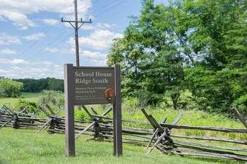  Mid-shot of the sign for School House Ridge South. Behind the sign is a brown split rail fence that travels diagonally. The ground is grassy and there are trees in the right part of the frameâ€™s background. Power lines are in frame left. 