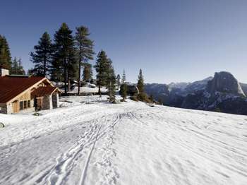 Ski track in the snow lead toward a wooden building with stone accents. Tall pine trees rise behind the building and a snow covered mountainous landscape, featuring Half Dome, can be seen to the right.