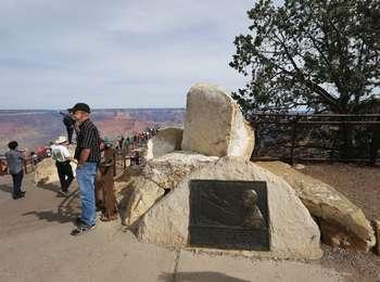 A rectangular metal plaque is mounted on a rough, uncut boulder of white limestone. Several more boulders litter the ground behind the one with the plaque. To the right of the plaque, a concrete walkway and series of steps lead down to a viewpoint looking out at the Grand Canyon landscape. Several people are standing or walking on the walkway and down the steps.