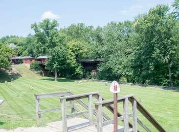  This image is taken from above on a wooden walkway. The wooden steps lead down to the grassy plot of land. The grassy plot stretches for the whole frame. In the background there are many trees. 