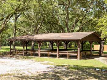 Wooden structure with wooden picnic tables underneath. Green field and trees surround the structure.