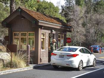 A park ranger steps out of a small brown kiosk to help a visitor in a white car.